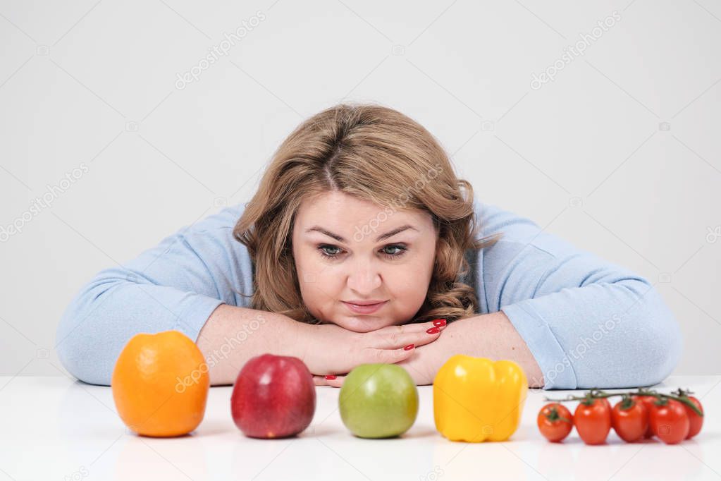 Young lush fat woman in casual blue clothes on a white background at the table, in front of her in a row laid out vegetables and fruits. Diet and proper nutrition.