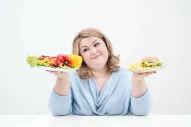 Holding bir sebze salatası ve fast food, hamburger ve patates kızartması bir tabak beyaz bir arka plan üzerinde rahat mavi giysili genç yemyeşil şişman kadın. Doğru beslenme ve diyet.