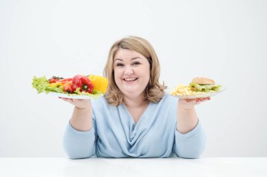 Holding bir sebze salatası ve fast food, hamburger ve patates kızartması bir tabak beyaz bir arka plan üzerinde rahat mavi giysili genç yemyeşil şişman kadın. Doğru beslenme ve diyet.