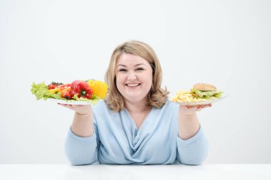 Holding bir sebze salatası ve fast food, hamburger ve patates kızartması bir tabak beyaz bir arka plan üzerinde rahat mavi giysili genç yemyeşil şişman kadın. Doğru beslenme ve diyet.