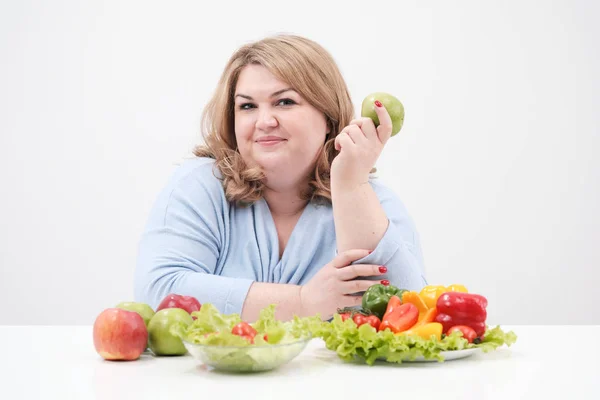 Jeune femme grasse courbée en vêtements bleus décontractés sur un fond blanc à la table et montre OK, légumes et fruits sont disposés devant elle dans une rangée. Alimentation et bonne nutrition . — Photo