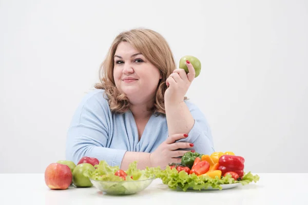 Jeune femme grasse courbée en vêtements bleus décontractés sur un fond blanc à la table et montre OK, légumes et fruits sont disposés devant elle dans une rangée. Alimentation et bonne nutrition . — Photo