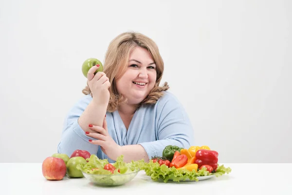 Молодая пышная толстая женщина в повседневной голубой одежде на белом фоне за столом и показывает ОК, овощи и фрукты выкладываются перед ней в ряд. Диета и правильное питание . — стоковое фото