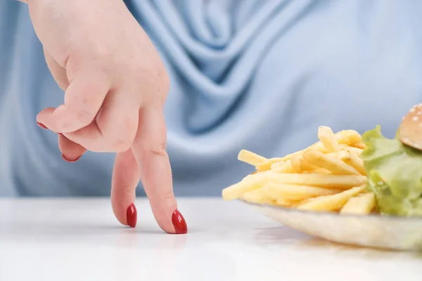 Los dedos de una joven exuberante mujer gorda en ropa azul casual sobre un fondo blanco, la elección entre la comida saludable y la comida rápida, concepto. Dieta y nutrición adecuada . — Foto de Stock