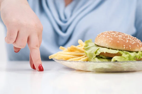 Los dedos de una joven exuberante mujer gorda en ropa azul casual sobre un fondo blanco, la elección entre la comida saludable y la comida rápida, concepto. Dieta y nutrición adecuada . — Foto de Stock