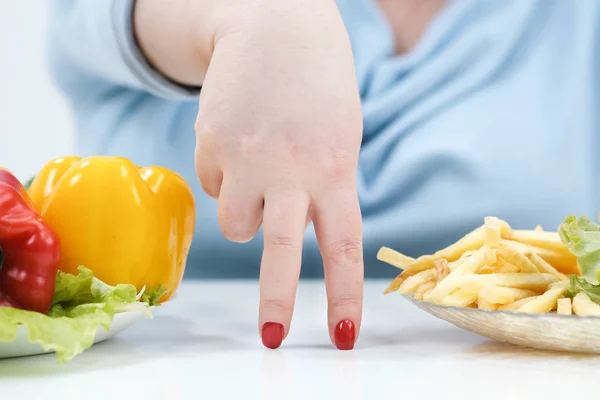 Dedos de uma jovem mulher gorda exuberante em roupas azuis casuais em um fundo branco, a escolha entre comida saudável e fast food, conceito. Dieta e nutrição adequada . — Fotografia de Stock