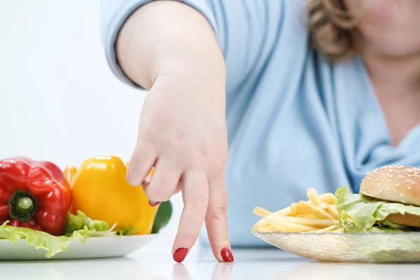 Los dedos de una joven exuberante mujer gorda en ropa azul casual sobre un fondo blanco, la elección entre la comida saludable y la comida rápida, concepto. Dieta y nutrición adecuada . — Foto de Stock