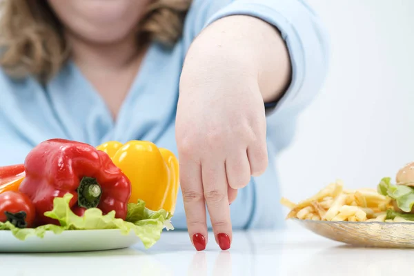 Los dedos de una joven exuberante mujer gorda en ropa azul casual sobre un fondo blanco, la elección entre la comida saludable y la comida rápida, concepto. Dieta y nutrición adecuada . — Foto de Stock