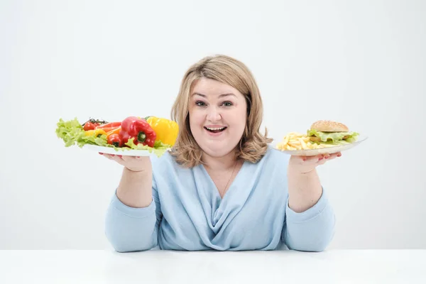 Jeune femme de graisse luxuriante dans des vêtements bleus décontractés sur un fond blanc tenant une salade de légumes et une assiette de restauration rapide, hamburger et frites. Alimentation et bonne nutrition . — Photo