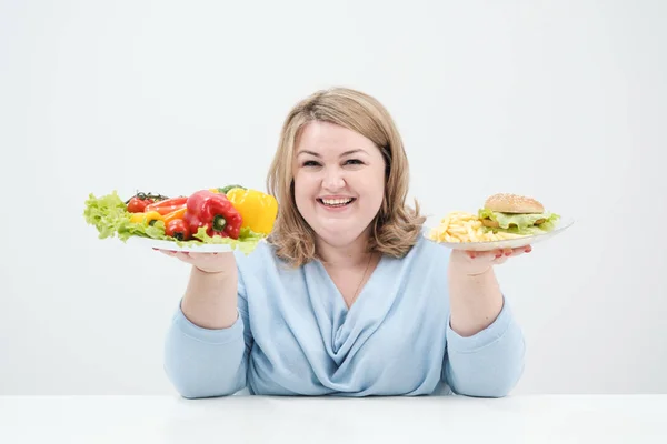 Молодая пышная толстая женщина в повседневной голубой одежде на белом фоне с овощным салатом и тарелкой фаст-фуда, гамбургером и картошкой фри. Диета и правильное питание . — стоковое фото