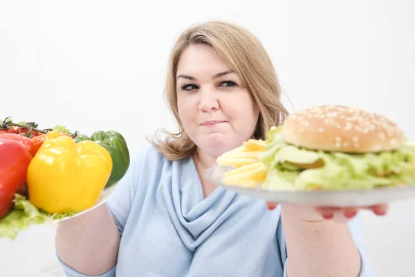 Jeune femme de graisse luxuriante dans des vêtements bleus décontractés sur un fond blanc tenant une salade de légumes et une assiette de restauration rapide, hamburger et frites. Alimentation et bonne nutrition . — Photo