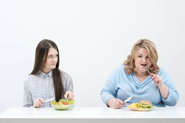 Szczupła dziewczyna zjada zdrową żywność, Gruba kobieta zjada szkodliwych fast food. Na białym tle, na temat diety i właściwego odżywiania. — Zdjęcie stockowe