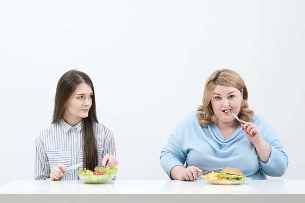 Chica delgada come comida saludable, mujer gorda come comida rápida dañina. Sobre un fondo blanco, el tema de la dieta y la nutrición adecuada . — Foto de Stock