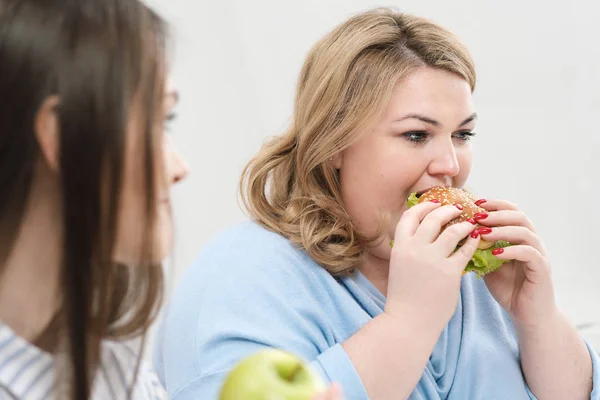 Szczupła dziewczyna zjada zdrową żywność, Gruba kobieta zjada szkodliwych fast food. Na białym tle, na temat diety i właściwego odżywiania. — Zdjęcie stockowe