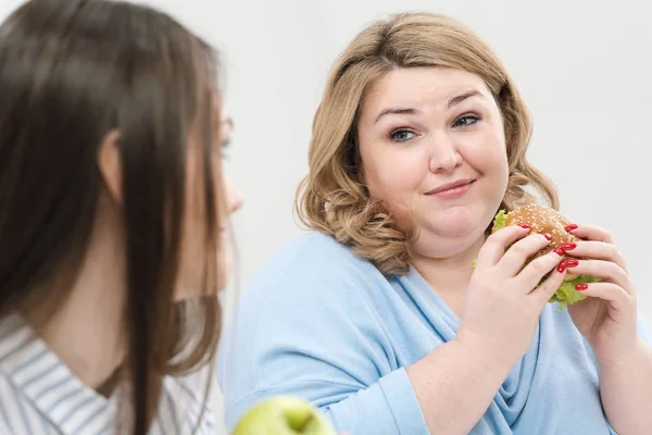 Chica delgada come comida saludable, mujer gorda come comida rápida dañina. Sobre un fondo blanco, el tema de la dieta y la nutrición adecuada . — Foto de Stock