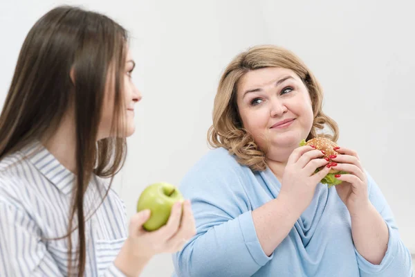 Струнка дівчина їсть здорову їжу, товста жінка їсть шкідливу фаст-фуду. На білому тлі тема дієти і правильного харчування . — стокове фото