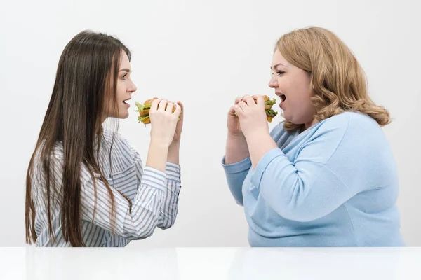 Dos chicas, delgadas y gordas, rubias y morenas, comen hamburguesas. Sobre un fondo blanco, el tema de la dieta y la nutrición adecuada . — Foto de Stock