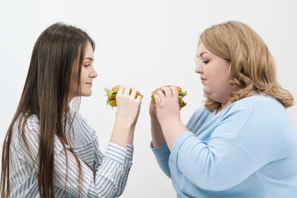 Duas meninas, magro e gordo, loira e morena, comer hambúrgueres. Em um fundo branco, o tema da dieta e nutrição adequada . — Fotografia de Stock