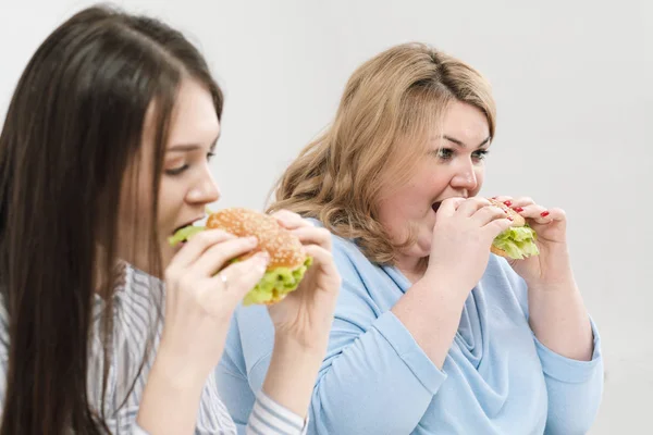 Duas meninas, magro e gordo, loira e morena, comer hambúrgueres. Em um fundo branco, o tema da dieta e nutrição adequada . — Fotografia de Stock