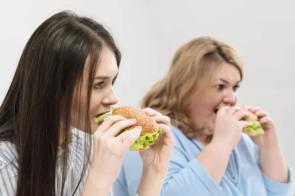 Duas meninas, magro e gordo, loira e morena, comer hambúrgueres. Em um fundo branco, o tema da dieta e nutrição adequada . — Fotografia de Stock