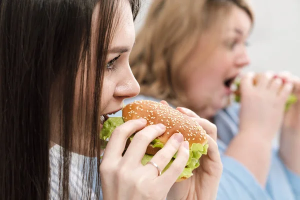 Dos chicas, delgadas y gordas, rubias y morenas, comen hamburguesas. Sobre un fondo blanco, el tema de la dieta y la nutrición adecuada . — Foto de Stock