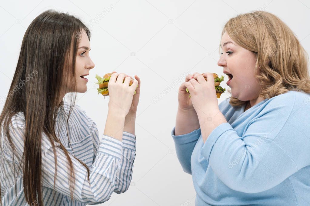 Two girls, slim and fat, blonde and brunette, eat hamburgers. On a white background, the theme of diet and proper nutrition.