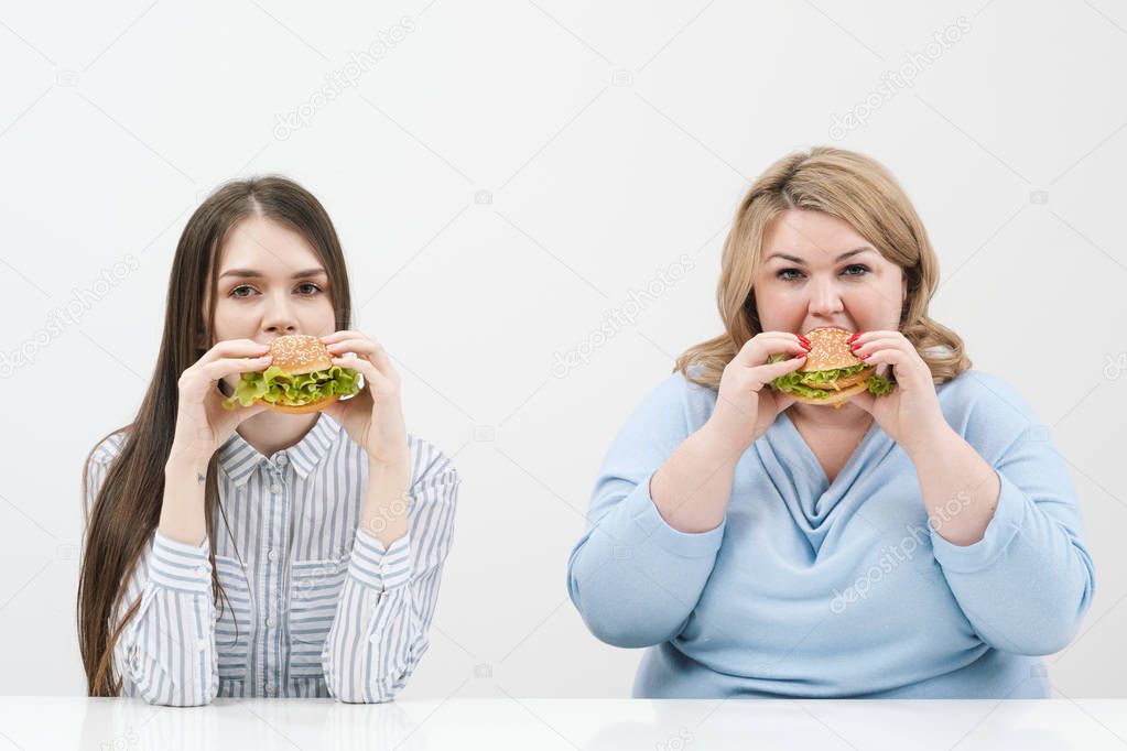 Two girls, slim and fat, blonde and brunette, eat hamburgers. On a white background, the theme of diet and proper nutrition.