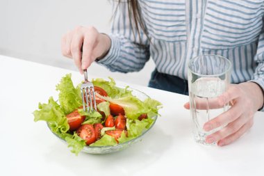 Beyaz bir arka plan üzerinde genç bir kız masada oturur ve domates ile yeşil salata çatalla yiyor, cam suyu içer. Sağlıklı gıda.