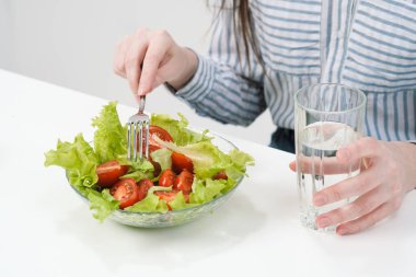 Beyaz bir arka plan üzerinde genç bir kız masada oturur ve domates ile yeşil salata çatalla yiyor, cam suyu içer. Sağlıklı gıda.