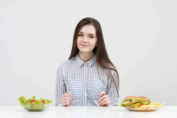 Štíhlá brunetka na bílém pozadí se rozhodne mezi desky fast food a hamburgery a zdravé jídlo salát. — Stock fotografie