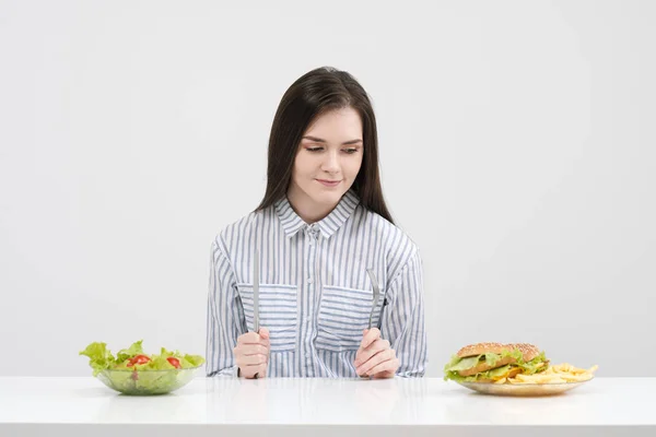 Štíhlá brunetka na bílém pozadí se rozhodne mezi desky fast food a hamburgery a zdravé jídlo salát. — Stock fotografie