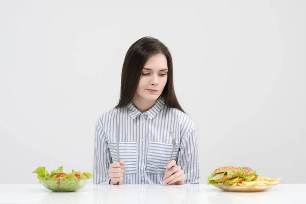 Štíhlá brunetka na bílém pozadí se rozhodne mezi desky fast food a hamburgery a zdravé jídlo salát. — Stock fotografie