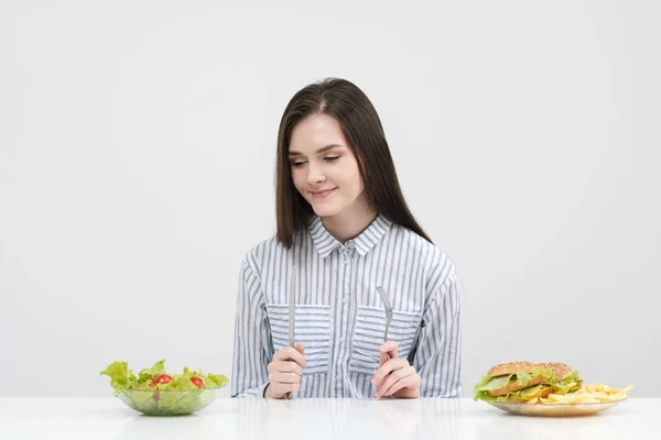 Mince fille brune sur un fond blanc choisit entre une assiette de restauration rapide et hamburgers et salade de nourriture saine . — Photo