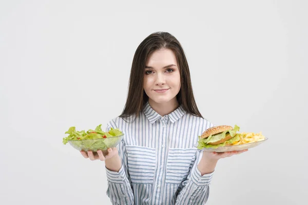 Mince fille brune sur un fond blanc choisit entre une assiette de restauration rapide et hamburgers et salade de nourriture saine . — Photo