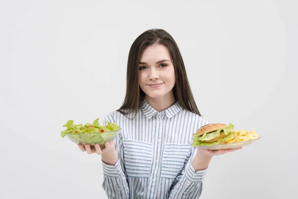 Štíhlá brunetka na bílém pozadí se rozhodne mezi desky fast food a hamburgery a zdravé jídlo salát. — Stock fotografie