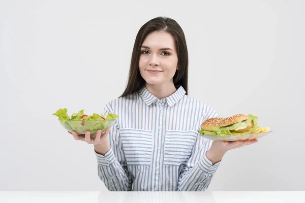 Струнка брюнетка дівчина на білому тлі вибирає між тарілкою фаст-фуду та гамбургерами та салатом здорової їжі . — стокове фото