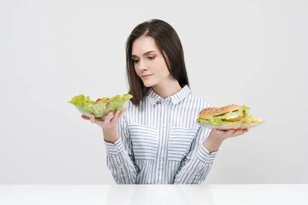 Chica morena delgada sobre un fondo blanco elige entre un plato de comida rápida y hamburguesas y ensalada de comida saludable . — Foto de Stock