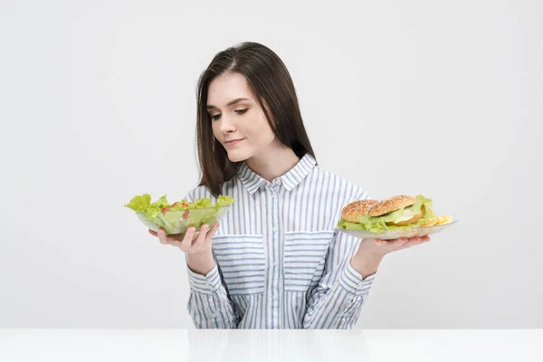 Mince fille brune sur un fond blanc choisit entre une assiette de restauration rapide et hamburgers et salade de nourriture saine . — Photo