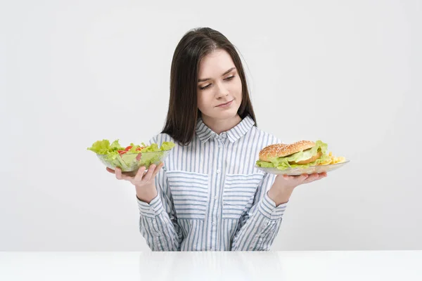 Menina morena esbelta em um fundo branco escolhe entre um prato de fast food e hambúrgueres e salada de comida saudável . — Fotografia de Stock