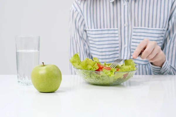 Une jeune fille sur fond blanc s'assoit à la table et mange une salade verte avec des tomates à la fourchette, boit de l'eau dans un verre. Aliments sains . — Photo