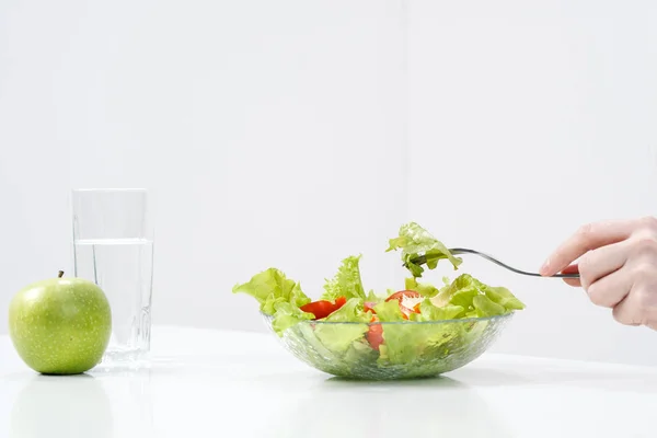 Mladá dívka na bílém pozadí sedí u stolu a jí zelený salát s rajčaty s vidličkou, pije vodu ze sklenice. Zdravé jídlo. — Stock fotografie