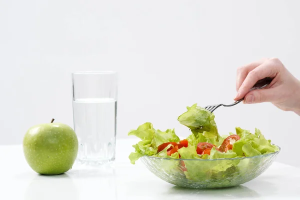 A young girl on a white background sits at the table and eats a green salad with tomatoes with a fork, drinks water from a glass. Healthy food. — Stock Photo, Image