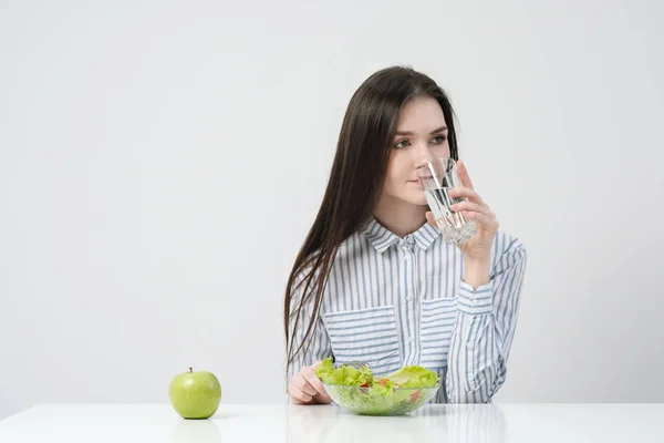 Une jeune fille sur fond blanc s'assoit à la table et mange une salade verte avec des tomates à la fourchette, boit de l'eau dans un verre. Aliments sains . — Photo