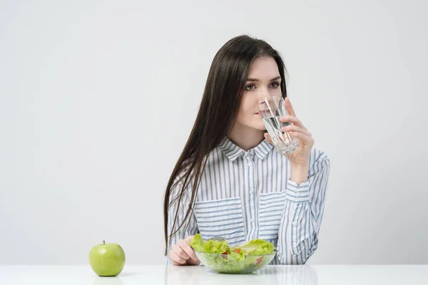 Une jeune fille sur fond blanc s'assoit à la table et mange une salade verte avec des tomates à la fourchette, boit de l'eau dans un verre. Aliments sains . — Photo