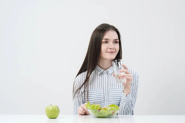 Une jeune fille sur fond blanc s'assoit à la table et mange une salade verte avec des tomates à la fourchette, boit de l'eau dans un verre. Aliments sains . — Photo