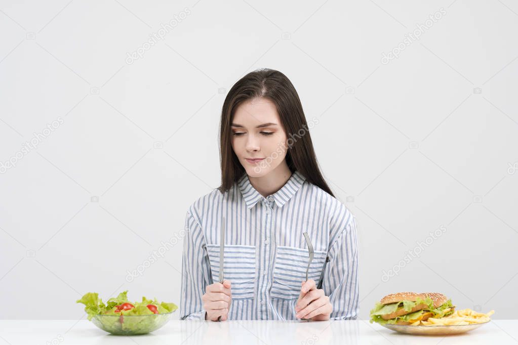Slender brunette girl on a white background chooses between a plate of fast food and hamburgers and healthy food salad.