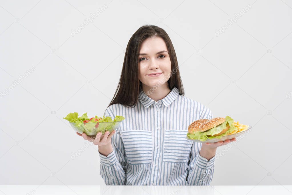 Slender brunette girl on a white background chooses between a plate of fast food and hamburgers and healthy food salad.