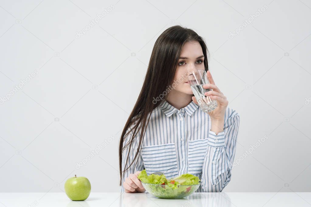 A young girl on a white background sits at the table and eats a green salad with tomatoes with a fork, drinks water from a glass. Healthy food.