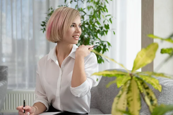 Ung vacker blond tjej med kort hår i en vit skjorta sitter på soffan i ljust på kontoret mot fönstret. Innehåller en bärbar dator och en smartphone. Pratar i telefon. — Stockfoto