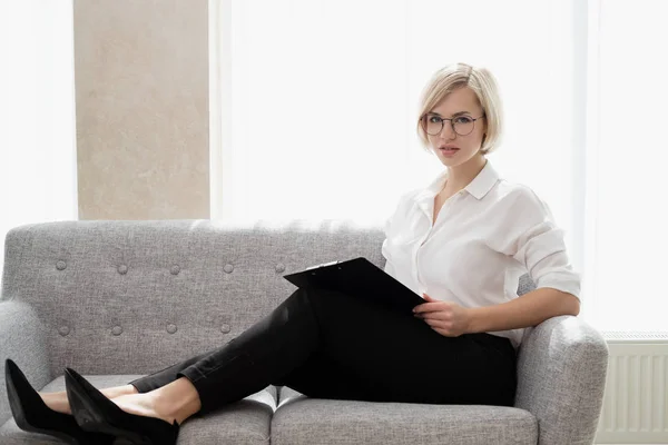 Ung vacker blond tjej med kort hår i en vit skjorta sitter på soffan i ljust på kontoret mot fönstret. Innehåller en bärbar dator och en smartphone. Pratar i telefon. — Stockfoto